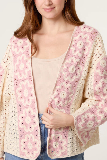 A woman wears a cream floral crochet cardigan with pink and white patterns, layered over a beige top and blue jeans, evoking vintage charm.