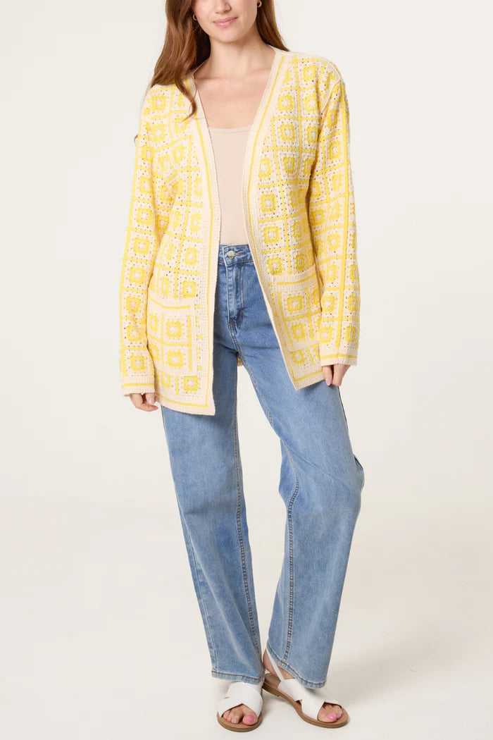 A woman in a yellow crocheted cardigan, light beige top, blue jeans, and white sandals stands against a plain background.