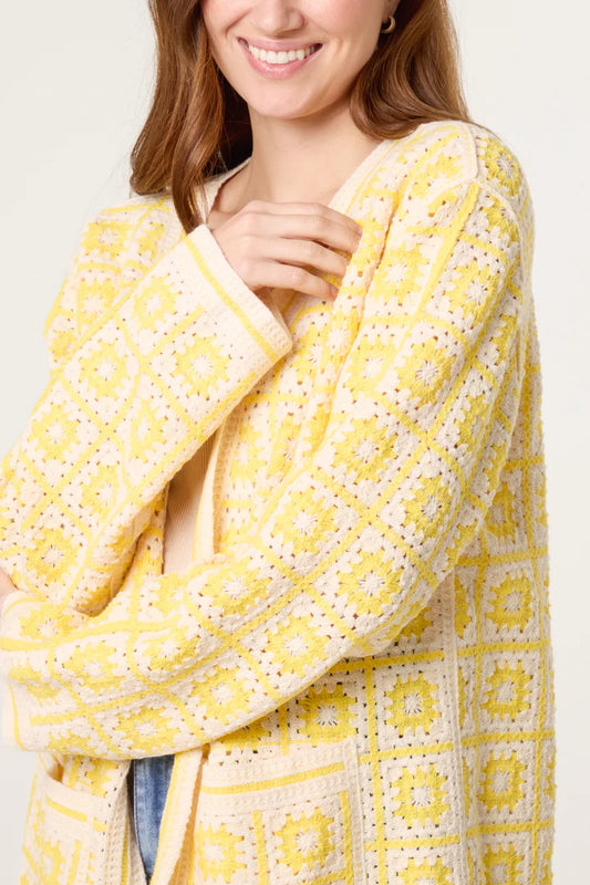 A woman wearing a yellow and white patchwork crochet cardigan with a square pattern stands with one arm crossed, smiling.