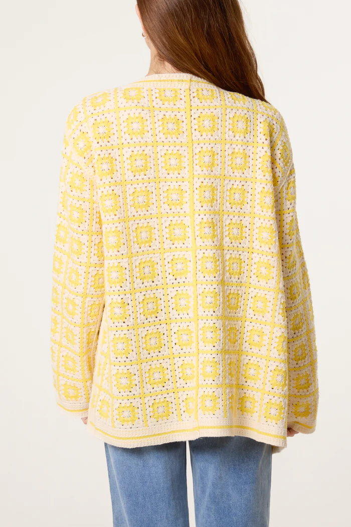 A person with long brown hair is seen from behind, wearing a yellow crochet cardigan featuring a square patchwork design, paired with light blue jeans.
