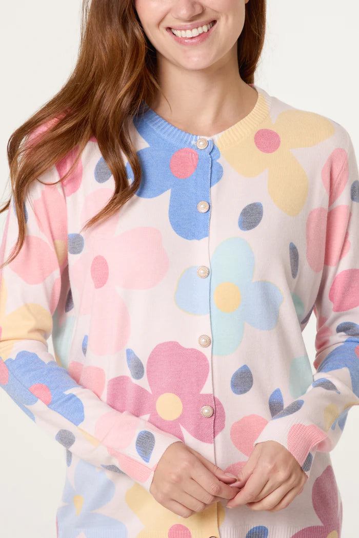 A woman wears a pastel floral cardigan with pearl buttons, made from vegan materials, smiling with her hands clasped together.