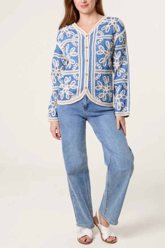 A woman wearing a vegan-friendly cardigan with white floral patterns, light blue jeans, and white sandals stands against a plain backdrop.