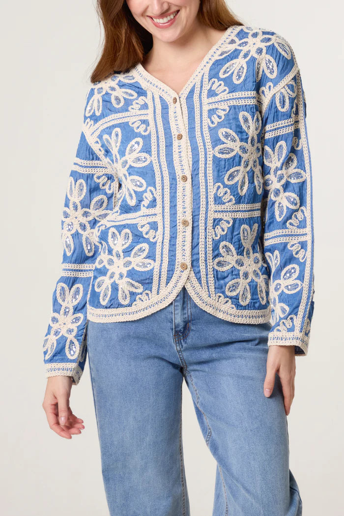 A woman wears a vegan-friendly blue cardigan adorned with white floral embroidery and light blue jeans, standing against a plain background.