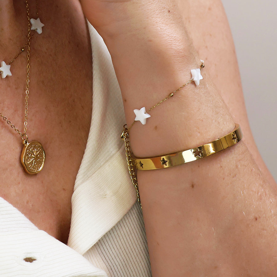A close-up of a person’s arm wearing a waterproof gold-plated star bracelet, a gold pendant necklace, and a fine chain with white star charms against a white shirt—perfect celestial jewellery.
