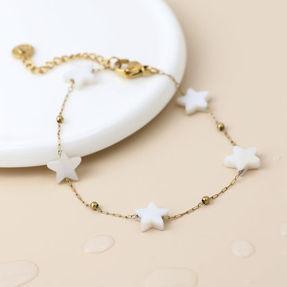 Delicate star bracelet with white star-shaped charms and small gold beads; this celestial jewellery piece is shown on a beige surface beside a white plate.