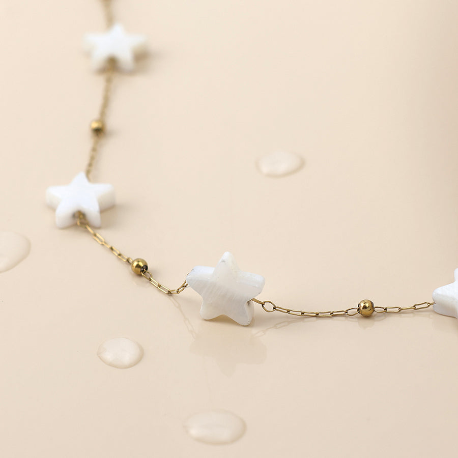 A gold chain necklace adorned with white star-shaped beads and small gold spheres, this celestial jewellery piece is displayed on a beige surface with droplets of water.