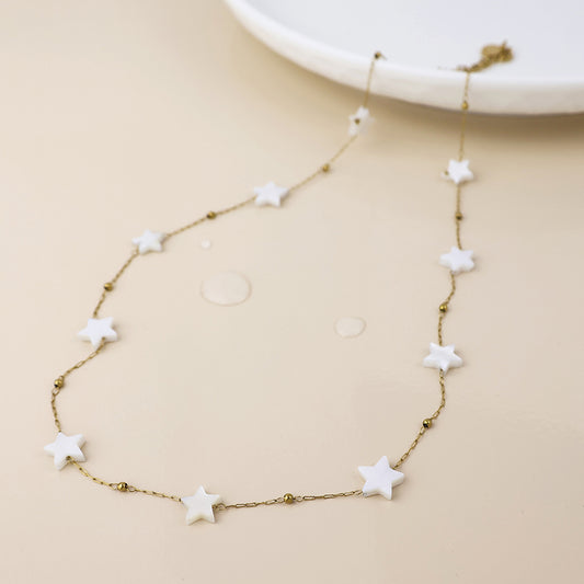 A tarnish-resistant gold star necklace featuring evenly spaced white star-shaped beads is shown on a light beige surface, with a white plate in the background.