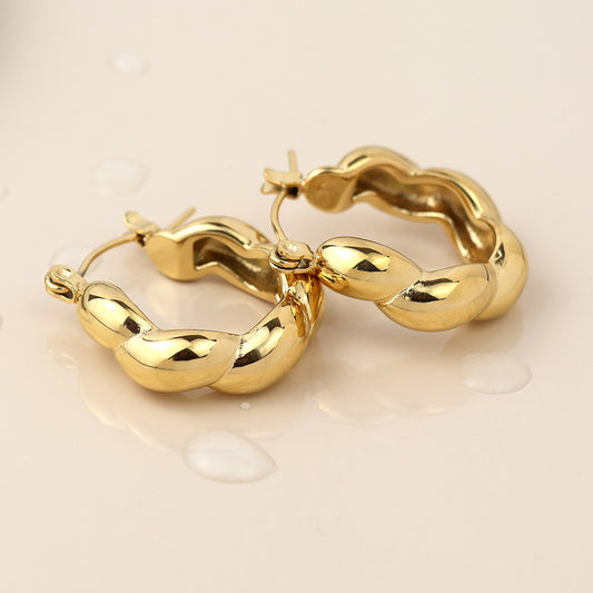 A pair of twist-design hoop earrings rests on a light surface with scattered water droplets, showcasing their elegant, tarnish-resistant finish.
