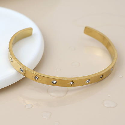 A gold open bangle bracelet featuring delicate star-shaped engravings and set with clear stones, shown on a beige surface with water droplets to emphasise its stylish and waterproof design.