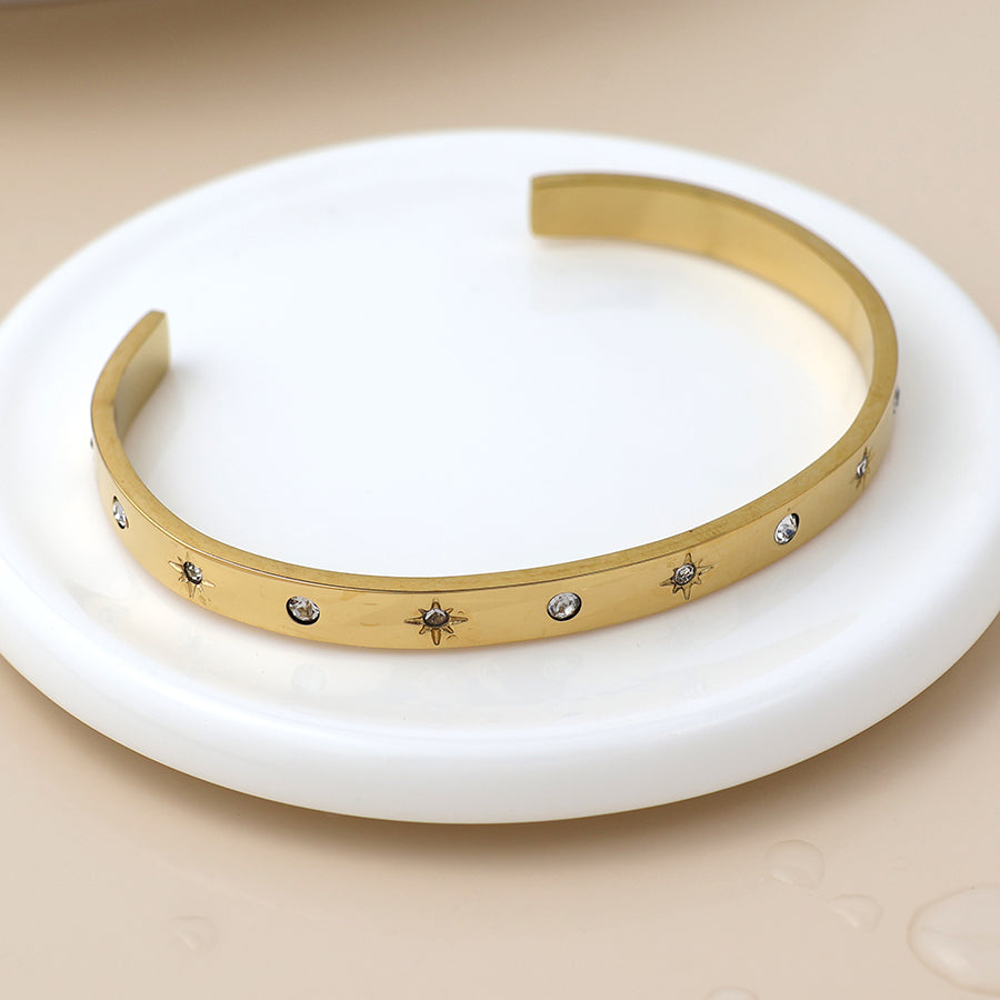 Gold open bangle featuring small engraved star motifs and set with clear stones, shown on a round white dish against a neutral backdrop.