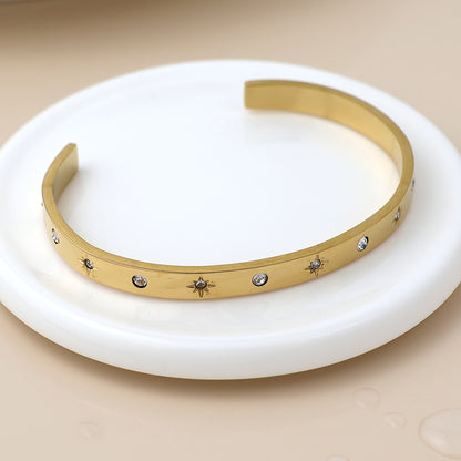 Gold open bangle featuring small engraved star motifs and set with clear stones, shown on a round white dish against a neutral backdrop.