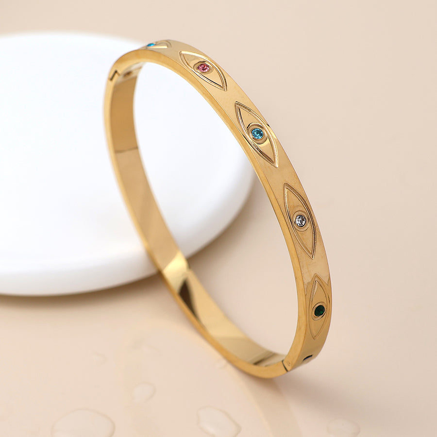 Gold bangle bracelet featuring oval engravings, each adorned with a small coloured gemstone, designed in an evil eye style and shown on a beige surface beside a round white object.