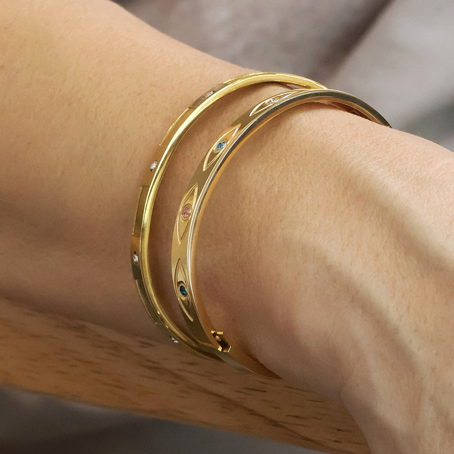 A close-up of a wrist adorned with two gold bangles: one plain, the other featuring small coloured gemstones and engraved designs, including a waterproof gold-plated bangle inspired by evil eye jewellery.