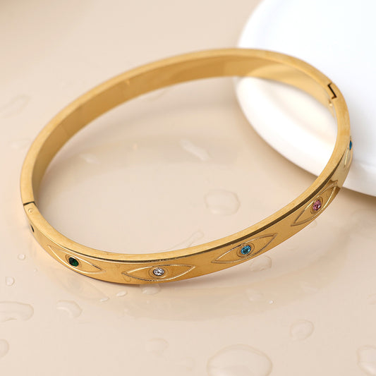 A waterproof gold bracelet adorned with engraved eye-shaped motifs and tiny multicoloured gemstones rests on a light surface, scattered with water droplets.