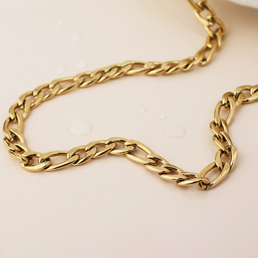 A close-up of a tarnish-resistant, waterproof gold-plated necklace featuring irregular links, resting on a light surface with several water droplets nearby.