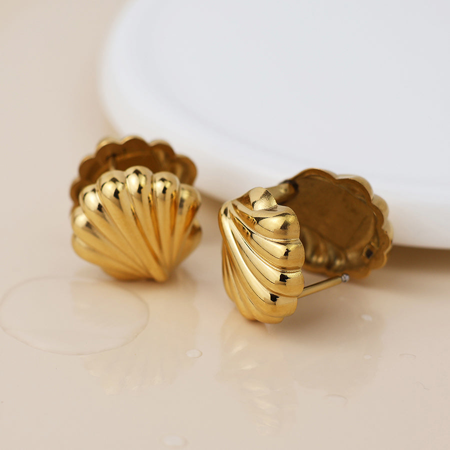 A pair of waterproof gold clam shell earrings displayed on a light surface beside a white ceramic plate.