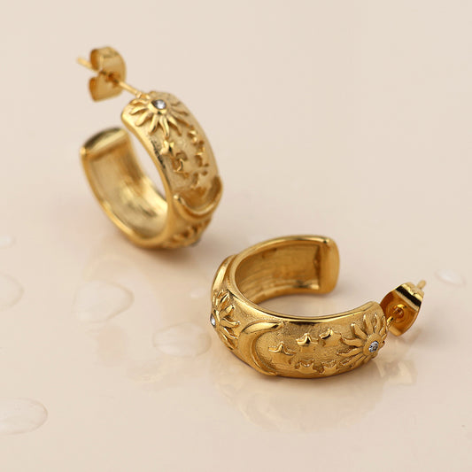 A pair of celestial hoop earrings with engraved floral patterns and small clear gemstones, displayed on a light surface with water droplets for a touch of cosmic jewellery elegance.