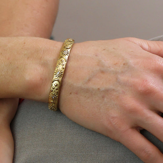 A gold-plated bracelet featuring a floral pattern, inspired by celestial bangle designs, is worn on a person’s wrist.