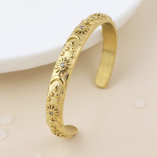 A gold-plated cuff bracelet with engraved moon, star, and flower motifs, adorned with small embedded gemstones—an elegant celestial bangle displayed on a light surface.