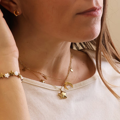 A woman in a white shirt layers celestial jewellery—a waterproof gold necklace with star and crescent moon charms, a double star necklace, a gold star bracelet, and a small gold hoop earring.