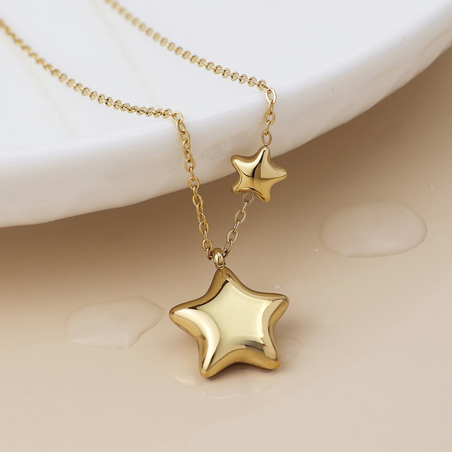 A celestial jewellery piece, this double star necklace showcases one large and one small gold pendant, elegantly displayed on a light-coloured surface beside a white dish.