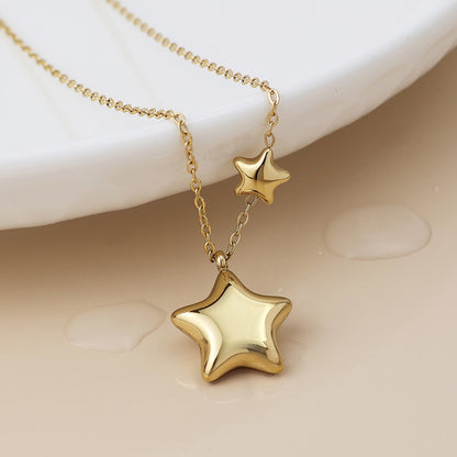 A celestial jewellery piece, this double star necklace showcases one large and one small gold pendant, elegantly displayed on a light-coloured surface beside a white dish.