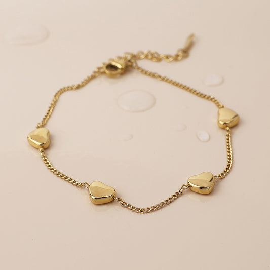 Delicate station bracelet with five gold-plated heart charms, shown on a light beige background with water droplets that emphasise its waterproof design.