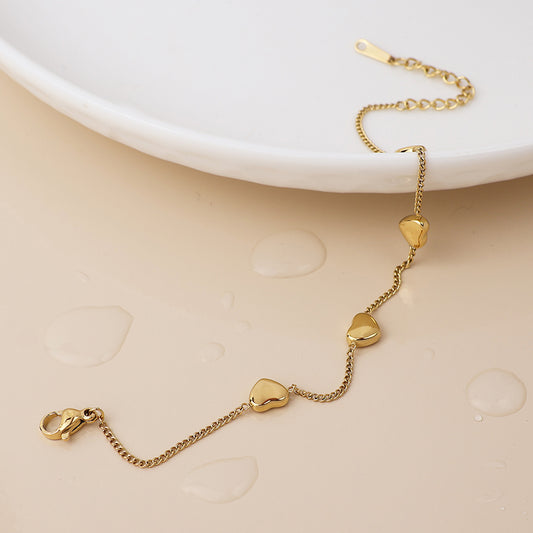 A gold-plated heart bracelet with three small charms lies on a beige surface with water droplets beside a white ceramic plate, emphasising its delicate, waterproof design.
