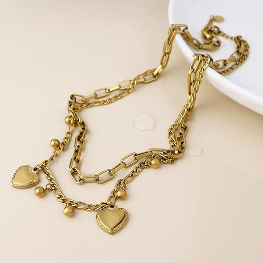 Waterproof gold-plated necklace with heart-shaped and round charms; this sturdy jewellery piece gleams on a pale surface beside the edge of a white dish.