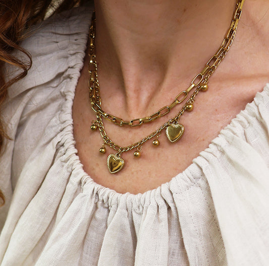 Close-up of a person wearing a layered hearts necklace with gold-plated heart and bead charms, matched with an off-white, ruched blouse. This hard-wearing jewellery is ideal for everyday elegance.