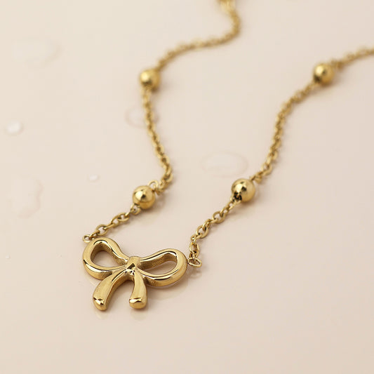 Gold-plated necklace with a dainty bow pendant and small round beads, shown against a light, neutral background.