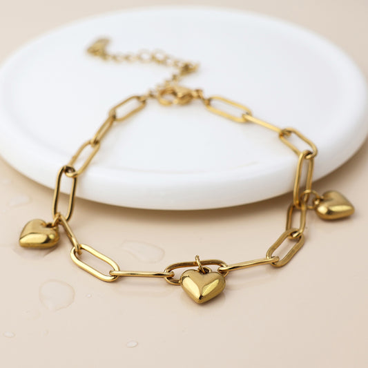 A gold-plated heart charm bracelet featuring three heart-shaped charms, presented on a round white dish.