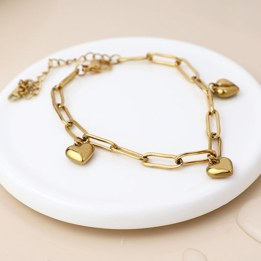 Gold-plated heart charm bracelet featuring rectangular links and three heart-shaped charms, arranged on a round white surface set against a beige background.