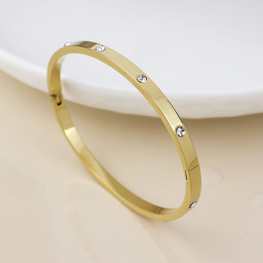 Gold-plated bangle bracelet adorned with round clear stones, this sparkling jewellery piece is shown on a light surface with a white dish in the background.