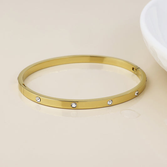 A gold-plated gem bangle decorated with small round clear stones, sparkling jewellery displayed on a light beige background beside a white dish.