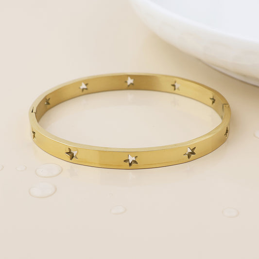 A waterproof gold-plated bangle featuring star-shaped cut-outs rests on a light beige surface beside a white dish, adding a stylish hint of celestial jewellery.