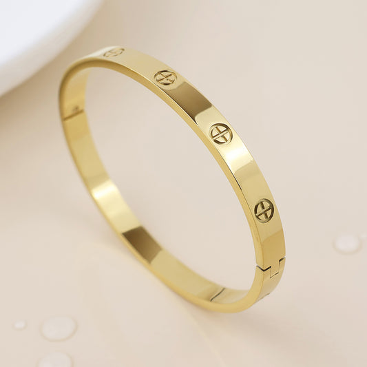 A waterproof gold-plated bangle bracelet featuring engraved screw motifs, shown on a light beige surface with several water droplets close by.
