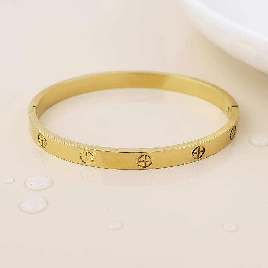 A waterproof, gold-plated bangle featuring screw motif engravings lies on a glossy beige surface beside water droplets, embodying fashionable bohemian jewellery style.