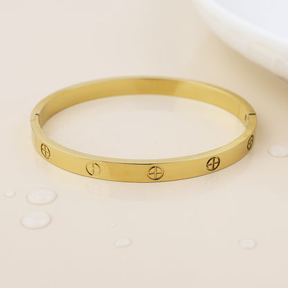 A waterproof, gold-plated bangle featuring screw motif engravings lies on a glossy beige surface beside water droplets, embodying fashionable bohemian jewellery style.