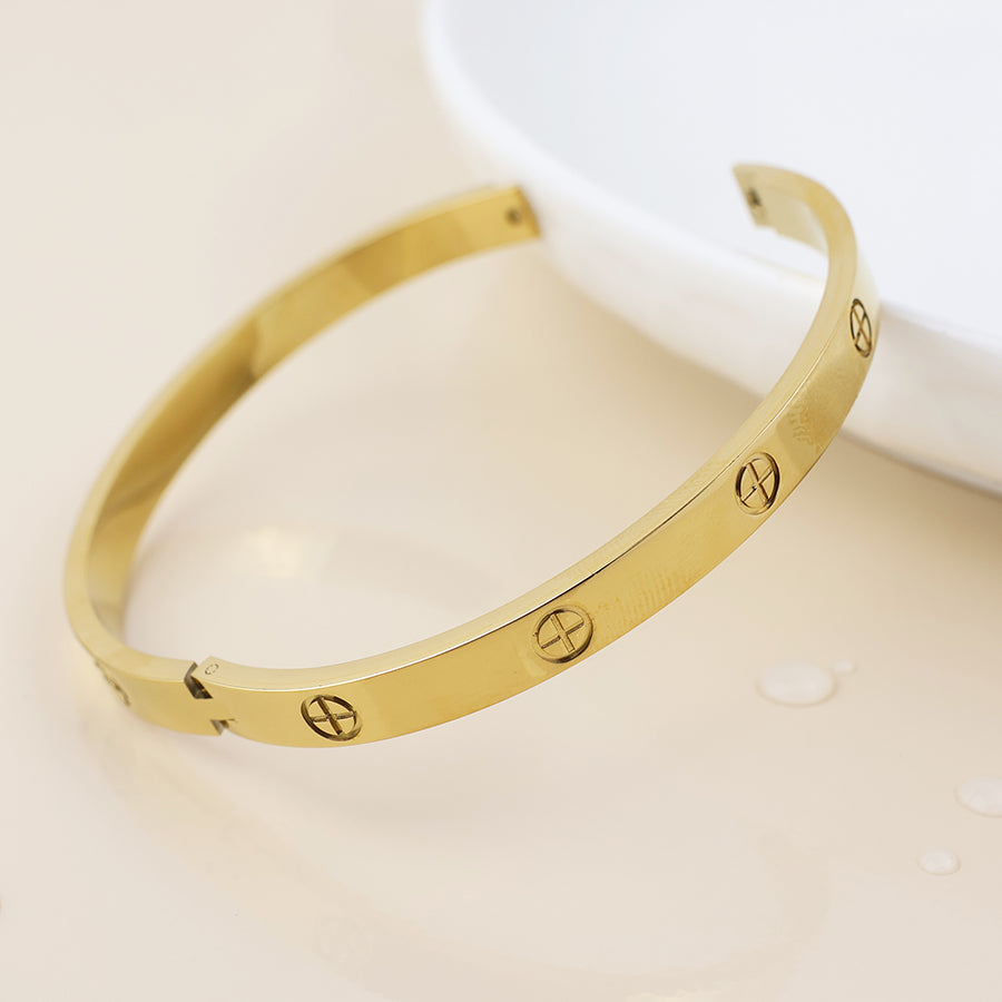 A waterproof gold-plated bangle with screw motifs lies on a cream-coloured surface beside a white dish, exemplifying elegant bohemian jewellery style.