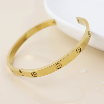 A waterproof gold-plated bangle with screw motifs lies on a cream-coloured surface beside a white dish, exemplifying elegant bohemian jewellery style.