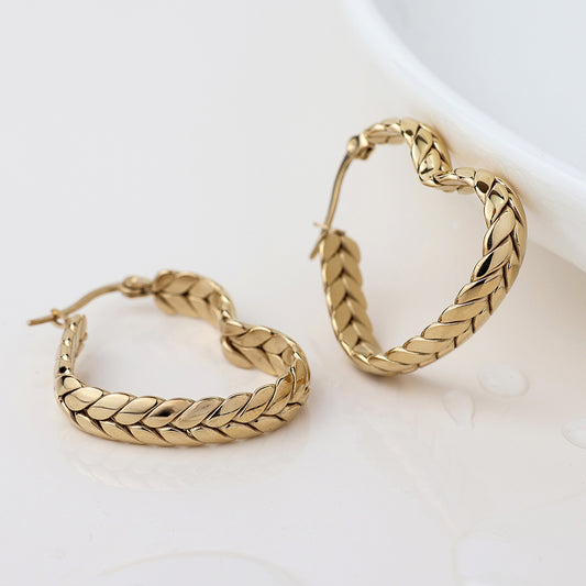 A pair of waterproof, gold-plated, braid-patterned heart hoop earrings rest on a white surface near a curved dish, ideal as statement earrings for any occasion.
