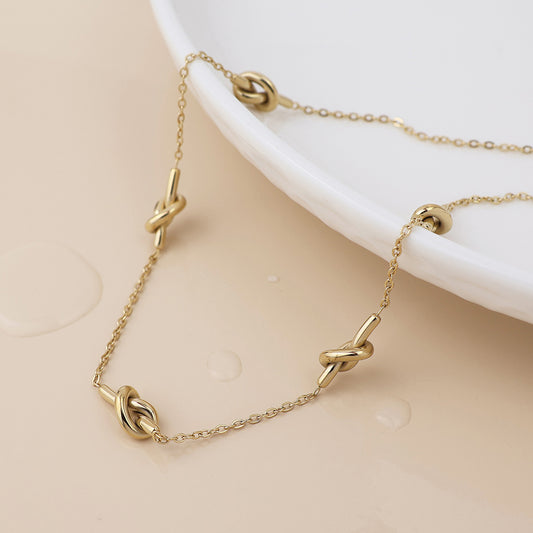 A waterproof gold-plated necklace featuring multiple knot-shaped links lies on the edge of a white ceramic dish, highlighted by nearby water droplets.