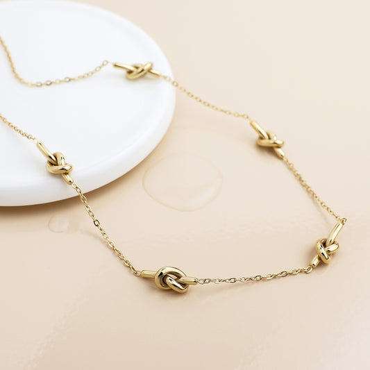 A waterproof, gold-plated necklace with elegant knot detailing, featuring several knot-shaped links, displayed on a light beige surface beside a round white dish.