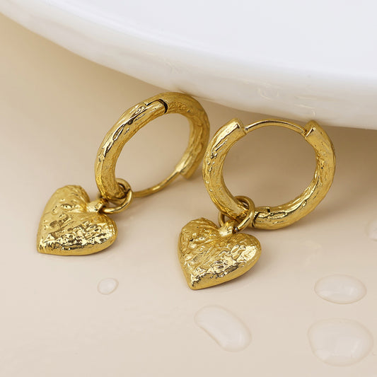 A pair of textured gold-plated hoops with dangling heart-shaped charms, displayed on a pale surface near drops of water—stylish heart earrings that catch every glimmer.