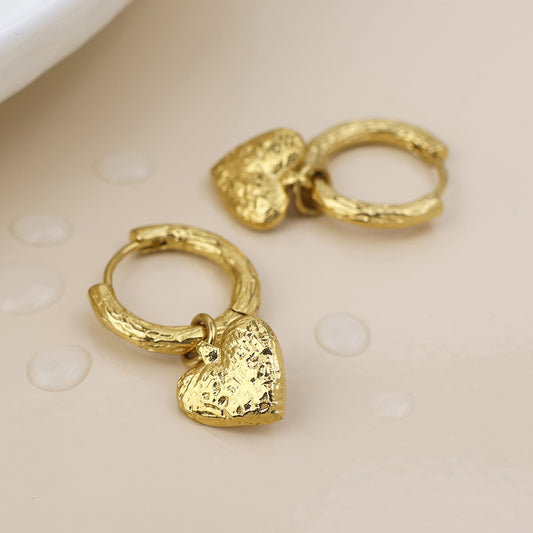 A pair of gold-plated hoops with heart-shaped pendants rests on a light beige surface, forming elegant heart earrings ideal for any occasion.