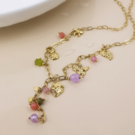 A waterproof gold-plated necklace adorned with fruit charms and colourful beads rests on a light surface beside a dish dotted with water droplets.
