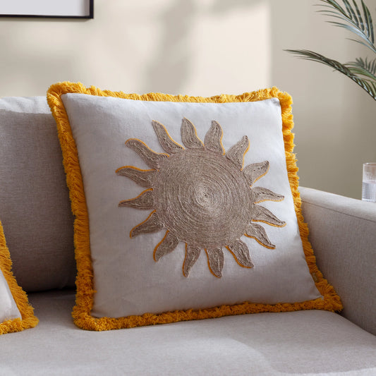 A sun-embroidered cushion featuring a sunflower design and fringe adorns a light-coloured sofa, next to a plant, adding a touch of elegant home decor.