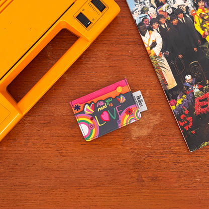 A colourful The Beatles cardholder with "All you need is love" text and rainbow designs rests on a wooden surface beside a yellow record player and a Beatles album cover.