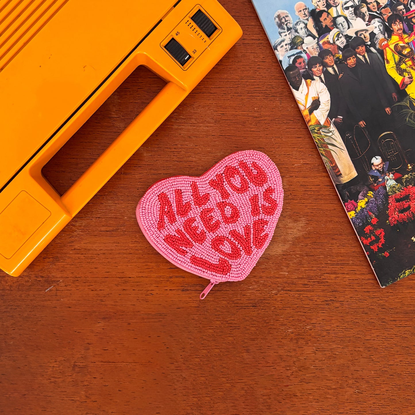 A music-themed accessory, this pink Beatles heart-shaped purse with ALL YOU NEED IS LOVE in red rests on a wooden surface beside an orange device and a colourful album cover.